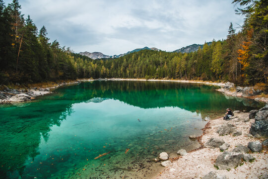 View of the serene turquoise lake reflects the dense forest and distant mountains under a cloudy sky in Eibsee, Garmisch-Partenkirchen, Bavaria, Germany.