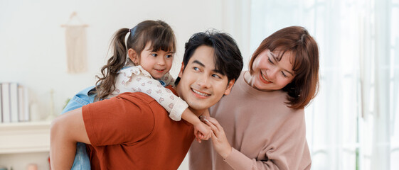 Happy Asian father giving daughter a piggyback ride at home while mother smiles at them in a cozy living room during a fun family moment