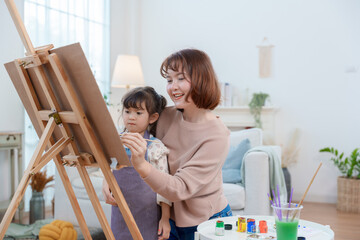 A happy Asian mother and her young daughter smiling while painting on an easel at home, enjoying a creative art activity and family time.