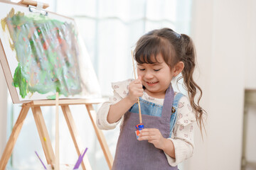 A happy little girl wearing an apron painting on canvas with a brush and easel at home. Creative child enjoying art lesson and preschool education development.