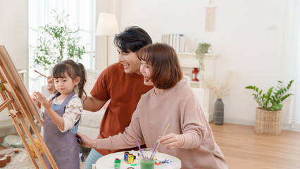 Happy Asian family with little daughter painting on canvas at home. Parents supporting child creative art development in living room.