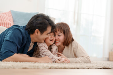 Happy Asian family lying on carpet at home father and mother hugging laughing daughter with love and warmth in cozy living room