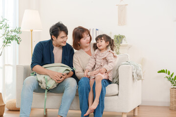 Happy young Asian family with little daughter sitting on sofa at home together father holding backpack preparing for school or travel