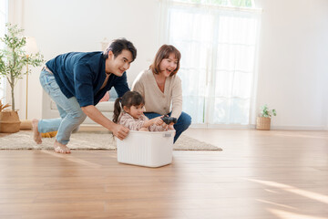 Happy Asian parents pushing their little daughter in a plastic basket while she holds a game controller at home Fun family bonding and creative childhood play in a bright modern living room