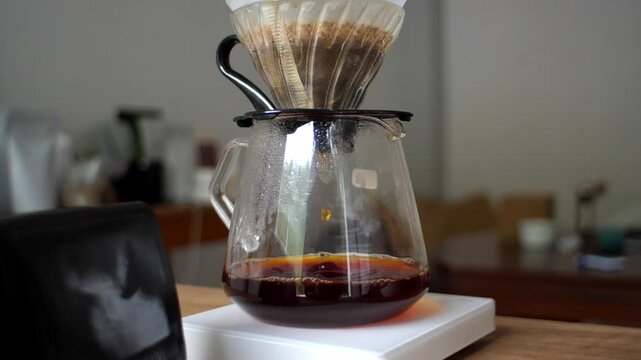 A glass server filled with freshly brewed filter coffee sits on a white scale on a wooden table. A coffee bean bag is visible in the foreground. Cozy morning atmosphere.