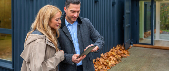 Portrait of architect explaining design concepts of modern modular house built from repurposed shipping containers in digital tablet to a satisfied client, with autumn leaves on the ground