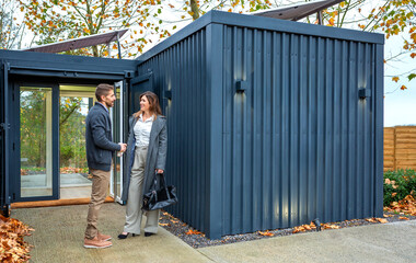 Professional couple conversing outside a modern, sustainable modular office built from repurposed shipping containers with solar panels and autumn leaves underfoot