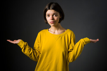 Young pretty girl in yellow sweater stands with outstretched arms and surprised facial expression, puzzled by the choice on gray background