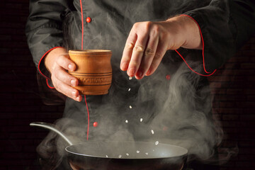 A cook stands in the kitchen, pouring coarse salt into a frying pan filled with smoke. He wears a...