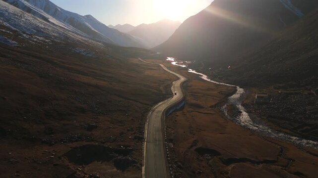 Aerial view of a winding road alongside a river, cutting through the rugged terrain with snow-capped mountains in the distance, Gittidas, Khyber Pakhtunkhwa, Pakistan.