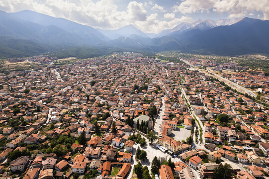 Aerial view of a dense urban sprawl nestled at the base of majestic, snow-capped mountains, a tapestry of red rooftops and green trees, Bansko, Blagoevgrad Province, Bulgaria.