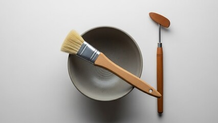 Artistic tools: a flat-bristled brush with a wooden handle and metal ferrule rests on a gray ceramic bowl, alongside a palette knife with a wooden handle and metal blade.