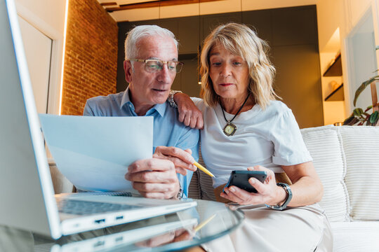 Senior couple calculating household finances and budgeting expenses at home, managing retirement savings and paying bills with a laptop and calculator for financial planning