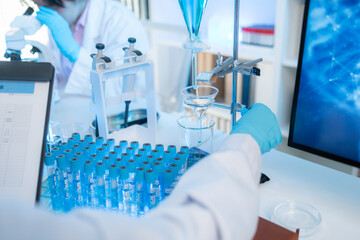 Two female scientists in white lab coats working together in a bright laboratory with microscopes, glassware, and blue test tubes, representing teamwork and modern scientific research environment.