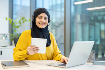Happy muslim businesswoman in hijab and headset multitasking with tablet and laptop in a modern office, smiling confidently during remote customer service or virtual meeting