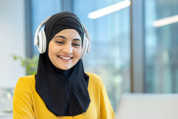 Young muslim woman wearing a hijab and white headphones, happily engaging with her laptop in a modern office environment, symbolizing connection, communication, and professional work ethic