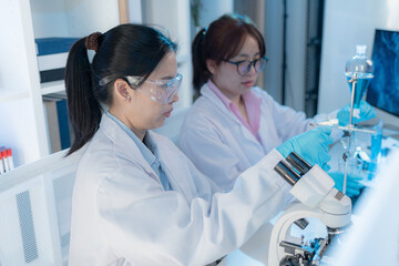 Two female scientists in white lab coats working together in a bright laboratory with microscopes, glassware, and blue test tubes, representing teamwork and modern scientific research environment.