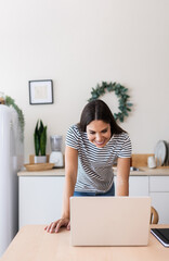 Young happy woman using laptop computer in the kitchen. Motivated female teleworking from home in a modern interior. Remote work and business lifestyle concept. Copy space for text.