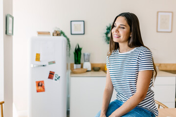 Young happy woman with long dark hair and wearing a striped t-shirt and jeans sitting confidently on kitchen table in a bright home. She is looking away and smiling. 