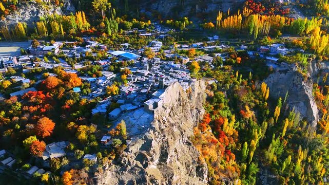 Aerial view of a town nestled on a cliffside, vibrant autumn foliage paints the landscape in warm hues, contrasting with the rugged terrain, Hunza, Gilgit Baltistan, Pakistan.