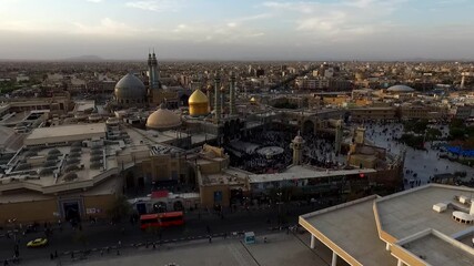 Drone panning over vast courtyard of Imam Reza Holy Shrine complex with golden dome prominent at dusk. Pilgrims and urban landscape in background ideal for faith and culture stock clips.