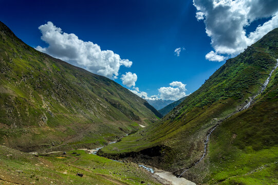 View of lush green mountains embracing a winding river under a vibrant blue sky dotted with fluffy white clouds, Noori Top, Khyber Pakhtunkhwa, Pakistan.