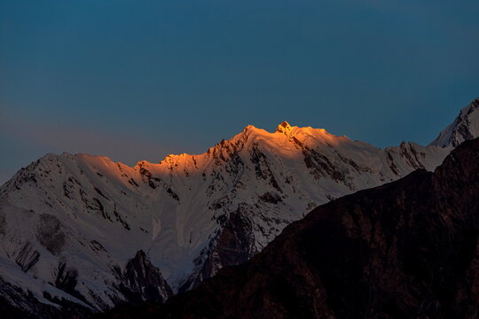 View of snow-capped peaks ablaze in the golden embrace of the setting sun, casting long shadows across the rugged terrain, Hunza, Gilgit Baltistan, Pakistan.