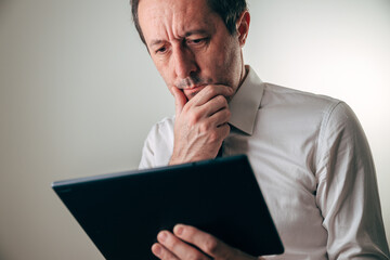 Businessman in white shirt using digital tablet to review financial report, looking worried and perplexed, expressing stress, uncertainty, and concern over business performance.