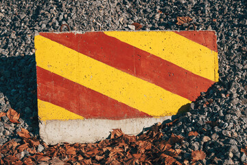 Concrete road barrier with yellow and red striped pattern standing in front of a large pile of gravel material.