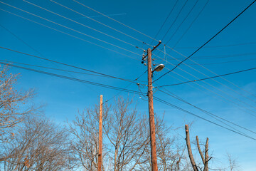 Low-angle view of weathered wooden electricity poles, tangled power lines, an old street lamp, and leafless winter trees against a bright clear blue sky.