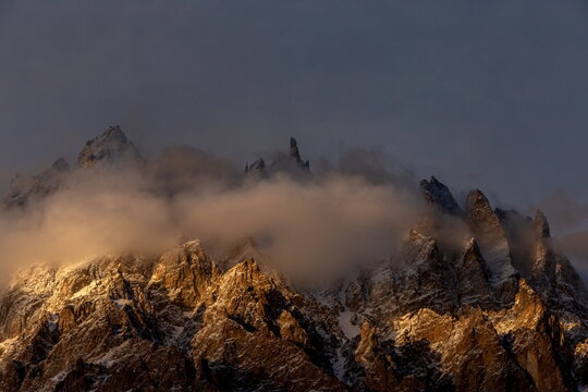 View of jagged, snow-dusted peaks pierce through swirling clouds, bathed in the warm glow of sunset along the Karakoram Highway, Passu, Gilgit Baltistan, Pakistan.
