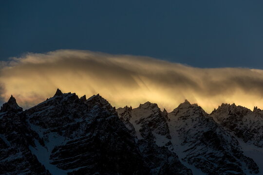 View of jagged, snow-dusted mountain peaks kissed by golden light beneath a swirling cloudscape against the deep blue sky, Passu, Gilgit Baltistan, Pakistan.