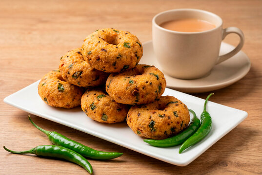 Delicious medu vada or savory lentil donuts served with hot indian chai tea and green chilies on a white rectangular plate on a wooden table