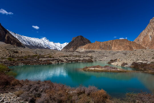 View of turquoise lake mirroring rugged, snow-capped mountains under an expansive blue sky, a serene vista in Passu, Gilgit Baltistan, Pakistan.