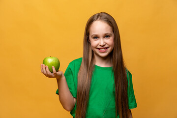 Happy smiling little girl in green t-shirt holding green apple and looking at camera isolated on yellow background. Proper nutrition. Child health. vegetarianism. High quality photo. Copy space