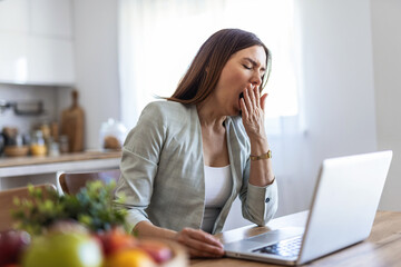 Bored young businesswoman yawning while going through paper and bills and working on a laptop at home. Tired female businessperson yawning while working from home.