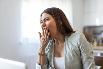Bored young businesswoman yawning while going through paper and bills and working on a laptop at home. Tired female businessperson yawning while working from home.