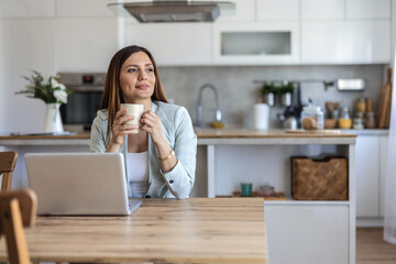 Dreamy young businesswoman using laptop and holding coffee cup enjoying freelance work sitting at desk indoors. Female entrepreneur having hot drink thinking looking aside at workplace.