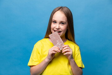 Cute little girl in yellow casual t-shirt eating chocolate bars on blue background, closeup. The concept of children's happiness and love for sweets. High quality photo. Copy space