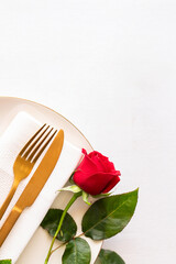 Place setting for a romantic dinner, with gold cutlery and a single long-stemmed red rose resting on a plate and napkin. The shot has a lot of white space for text or design.