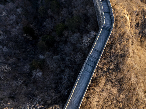 Aerial view of the Great Wall snaking through the rugged, forested terrain, a stone serpent dividing the dark woods from the sunlit slopes, Jingshanling, Beijing, China.