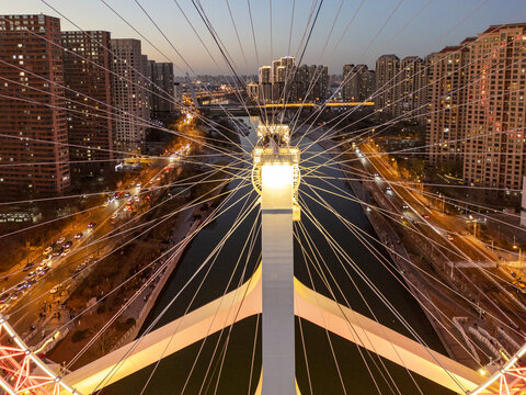 Aerial view of a modern bridge with intricate cable patterns over a dark river, contrasting with the warm glow of city lights at dusk, Tianjin, Tianjin, China.