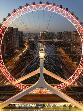 Aerial view of the radiant, crimson Tianjin Eye Ferris wheel arches gracefully over the Hai River, casting a vibrant glow against the dusk sky, Tianjin, Tianjin, China.