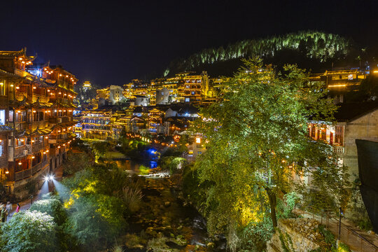 View of glowing buildings and bridges cast warm light over the dark river in a captivating panorama, Furong town, Hunan, China..
