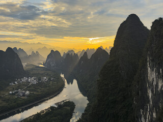 View of the Li River meanders through a landscape of sharp, dark karst peaks under a golden sunset, creating a dramatic contrast of light and shadow, Xingping, Guilin, China.