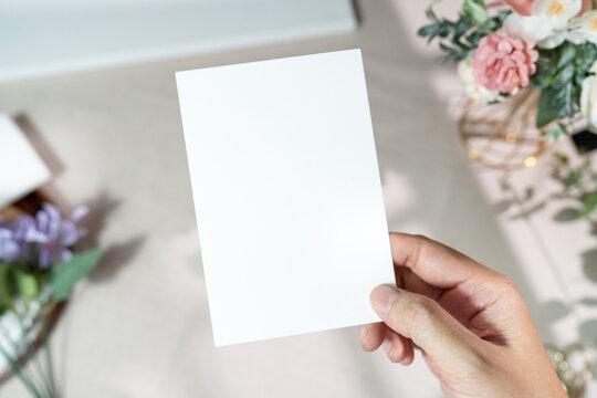 Mockup of a blank white card held by hand with soft blurred flowers, perfect for wedding or Valentine&rsquo;s greeting design, with clipping path.