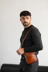 A stylish man in a dark grey crew neck sweater poses with a cognac leather crossbody bag against a minimalist white studio background.