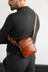 A stylish man in a dark grey crew neck sweater poses with a cognac leather crossbody bag against a minimalist white studio background.