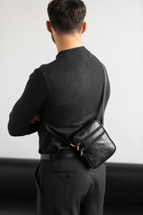 A stylish man in a dark grey crew neck sweater poses with a black leather crossbody bag against a minimalist white studio background.