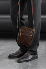 Stylish man in charcoal knitwear and trousers holds a dark brown vertical leather messenger bag against a white studio backdrop